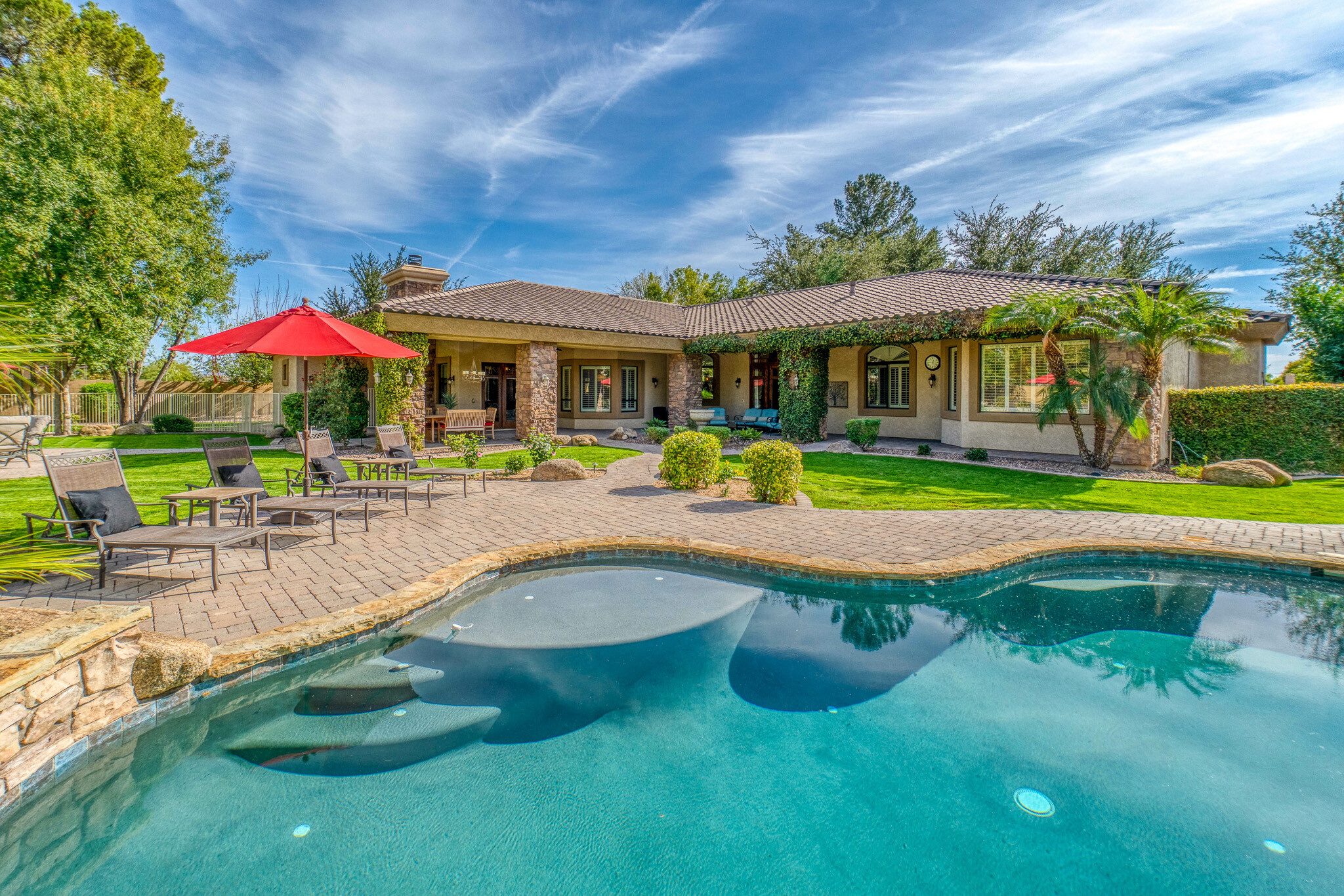 A backyard pool with many plants, seating, pool deck, and a red umbrella.