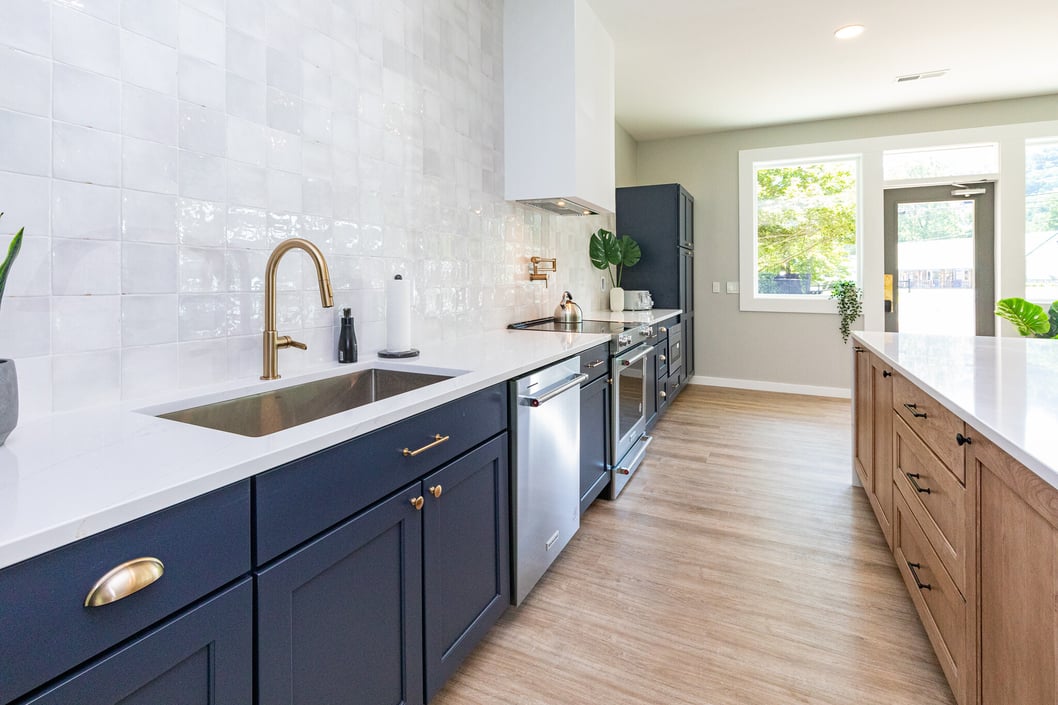 Bright and modern kitchen featuring navy blue lower cabinets with gold hardware, a white quartz countertop, and a farmhouse-style sink with a brushed gold faucet. A large island with natural wood cabinetry contrasts with the sleek white tile backsplash. Stainless steel appliances and large windows bring in natural light, creating an inviting and stylish space.