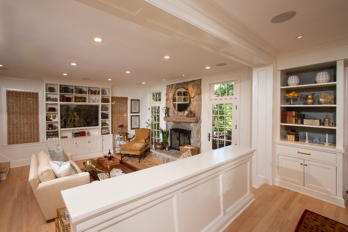 Family living room with stone fireplace, white wood shelving, overhead can lighting and a wooden ledge dividing the living room from the dining room