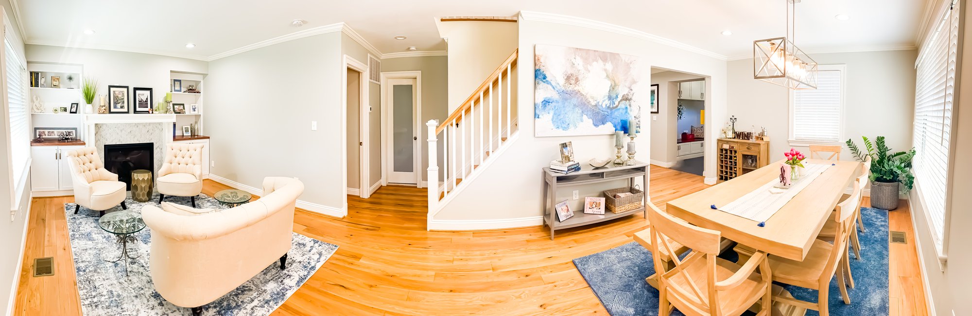 A combined living room and dining room.  Featuring cream colored sofa and arm chairs and wooden dining room table.  A staircase leads to the second floor from the center of the space. 