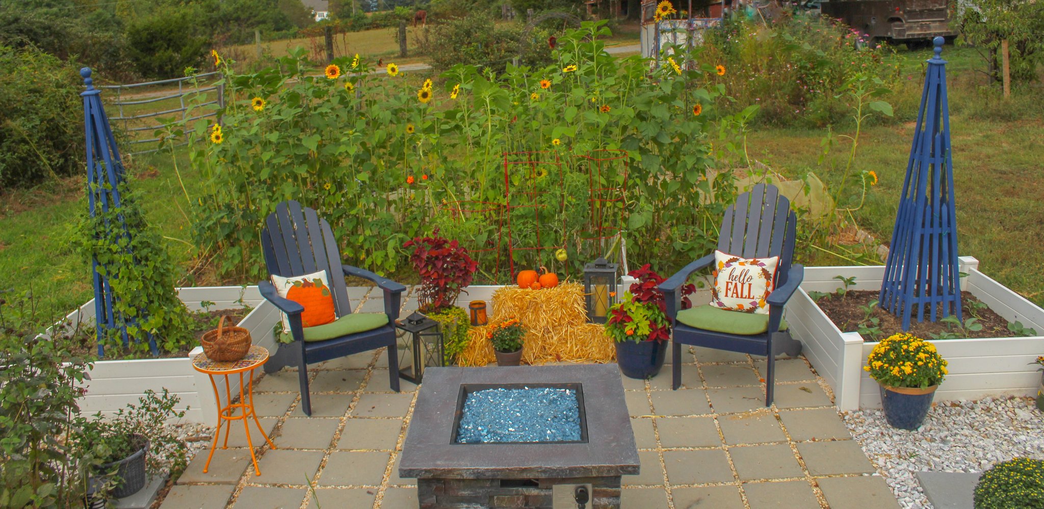 Fire pit in a backyard surrounded by chairs, sunflowers, pumpkins and bales of hay.